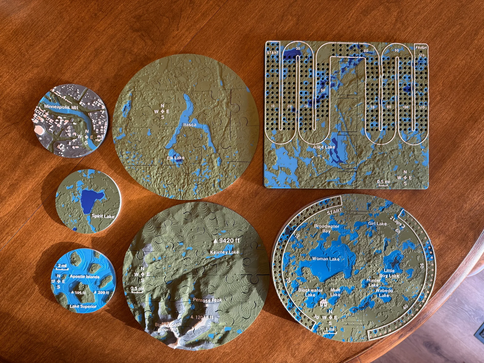 Full track and oval folding cribbage boards, two topographic jigsaw puzzles and three landscape magnets laid out together on a wooden table