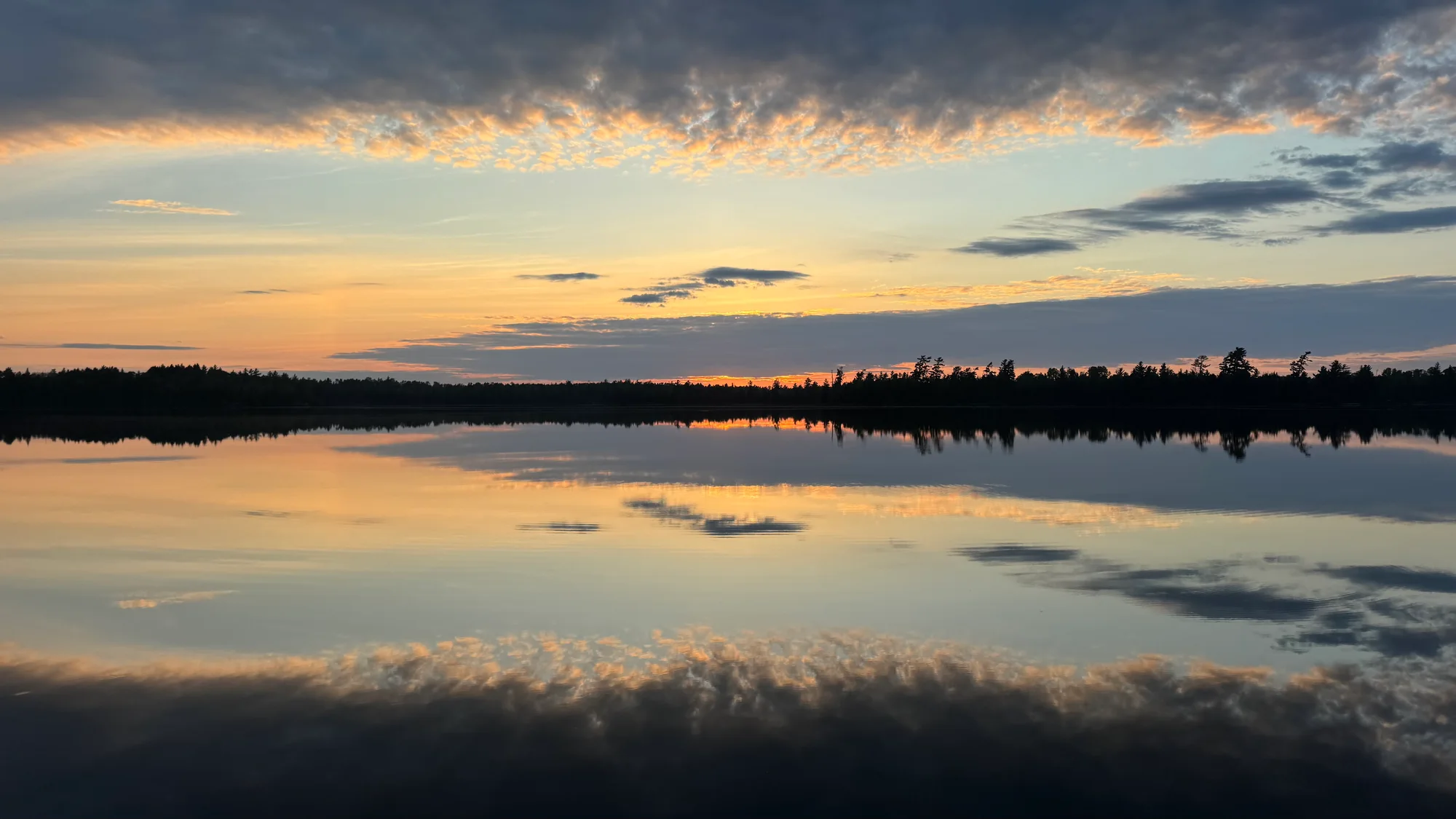 Gold and pink sunset clouds reflected across a calm lake and wooded shoreline
