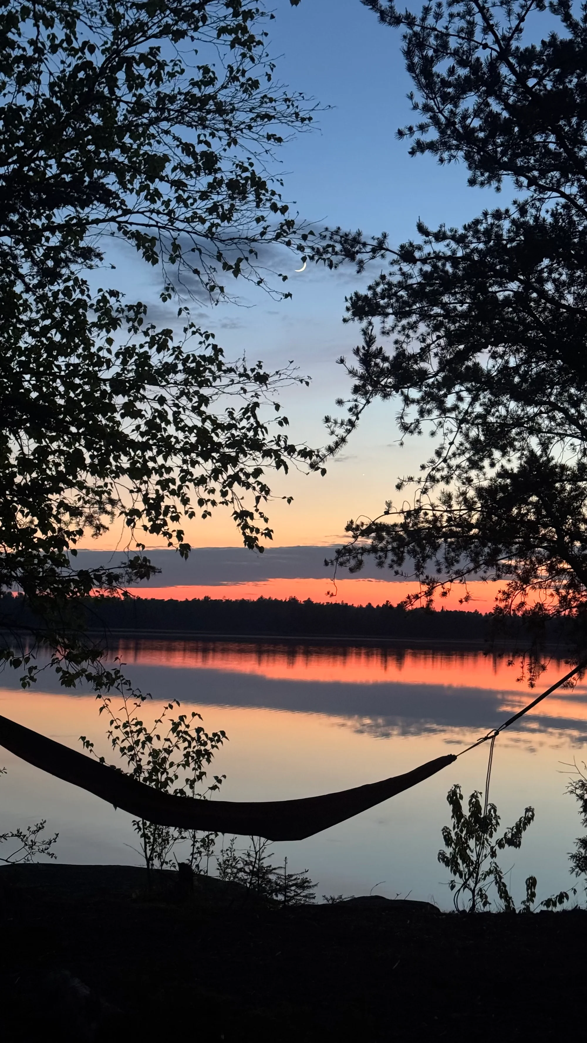 A hammock between trees beside a still lake, with a crescent moon above the sunset
