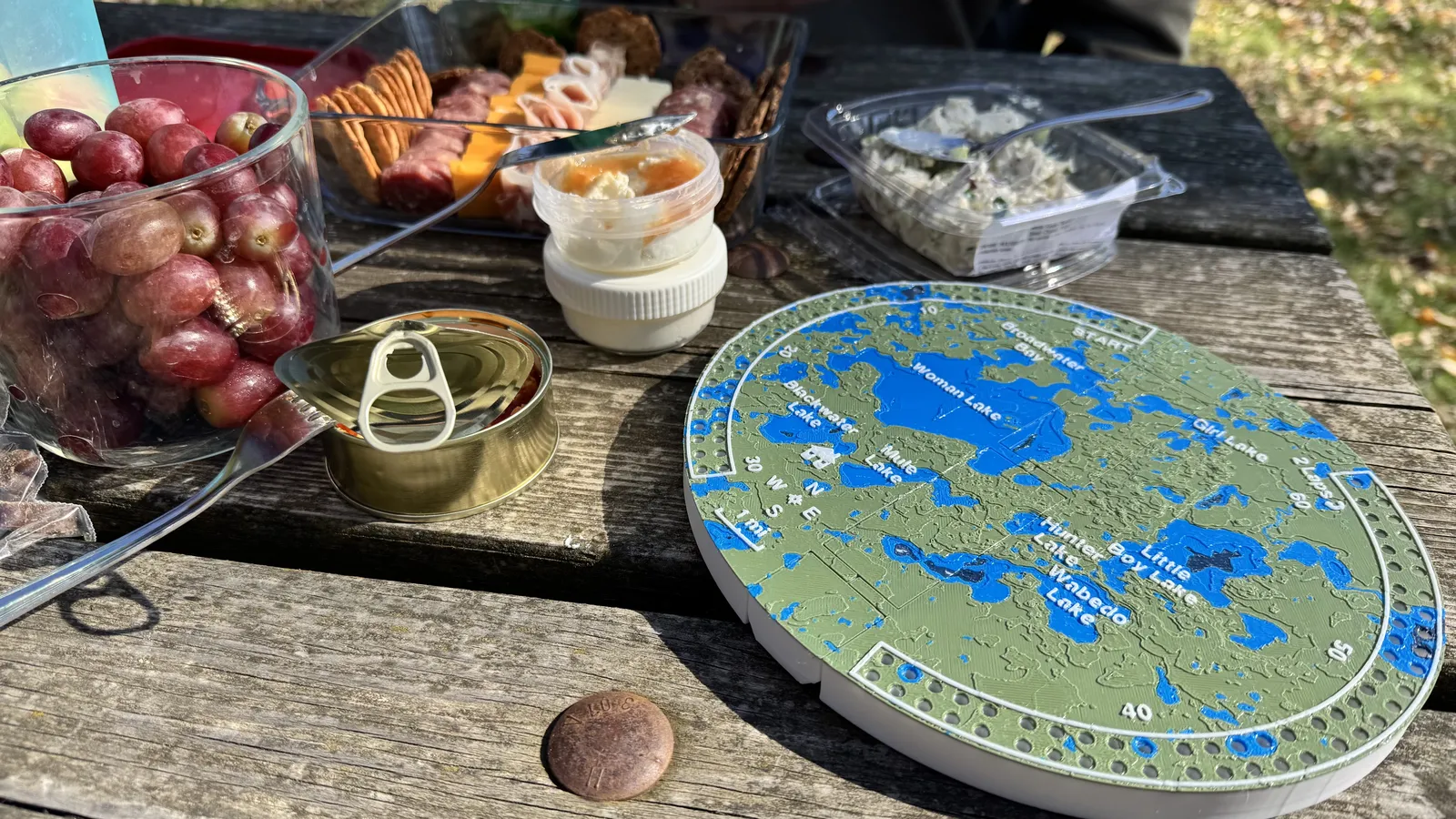 Open folding topographic cribbage board beside picnic food on a wooden picnic table